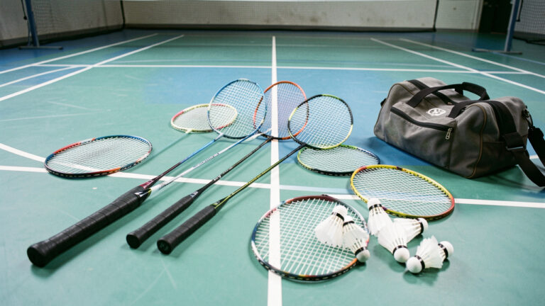 Plusieurs raquettes de badminton de styles variés disposées sur un terrain intérieur avec des volants, illustrant le choix d’un modèle adapté au niveau de jeu.